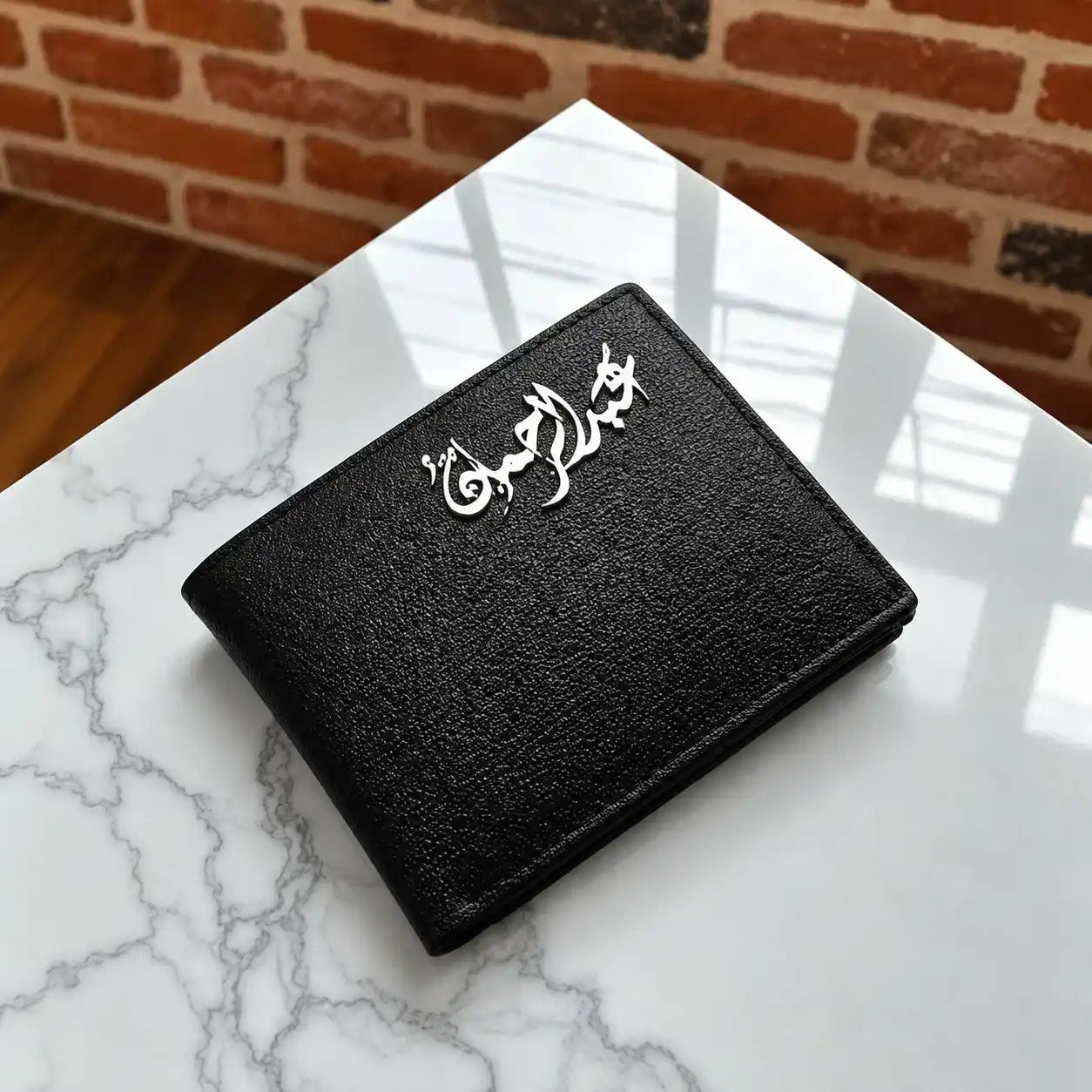 Black textured metallic wallet with silver customized Arabic calligraphy emblem on a white marble glass table.
