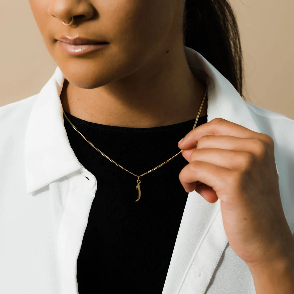 Gold Arabic initial letter charm necklace worn by a woman wearing a white collared shirt over a black top.