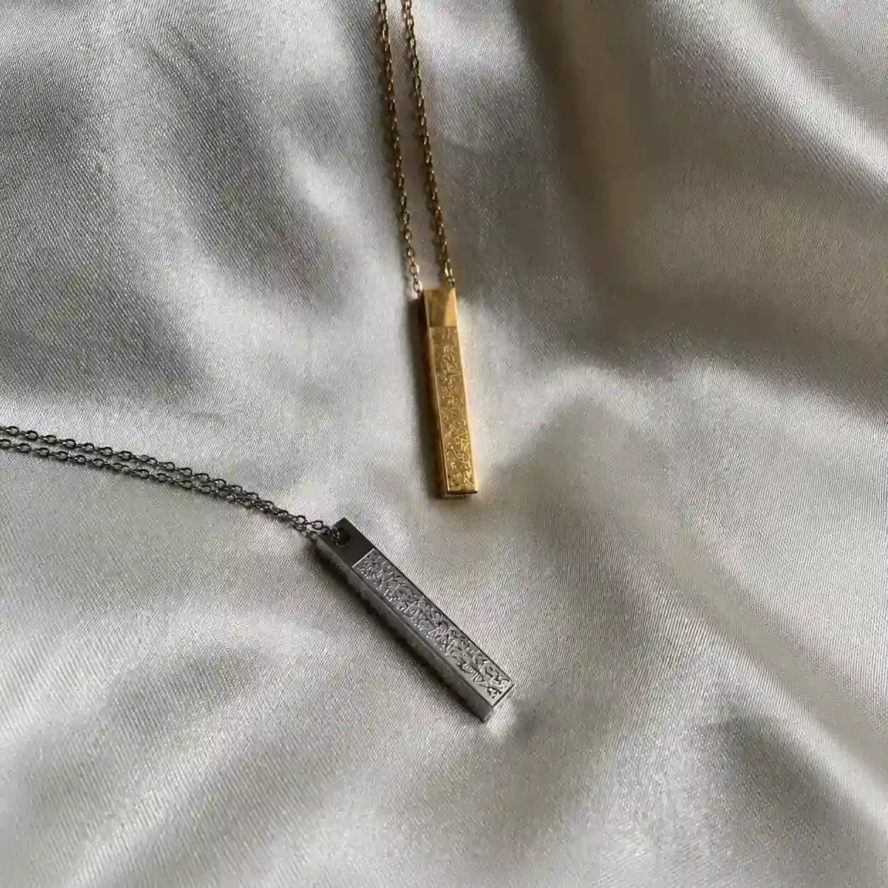 Gold and silver Ayat Ul Kursi bar pendant necklace with engraved Arabic calligraphy displayed on a white satin fabric background.