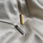 Gold and silver Ayat Ul Kursi bar pendant necklace with engraved Arabic calligraphy displayed on a white satin fabric background.