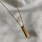 A gold bar necklace with a textured, engraved rectangular pendant on a delicate gold chain, displayed on a draped light gray fabric background.