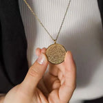 A close-up of a person holding a round gold pendant necklace with intricate Arabic calligraphy, wearing a white ribbed shirt and a black blazer.