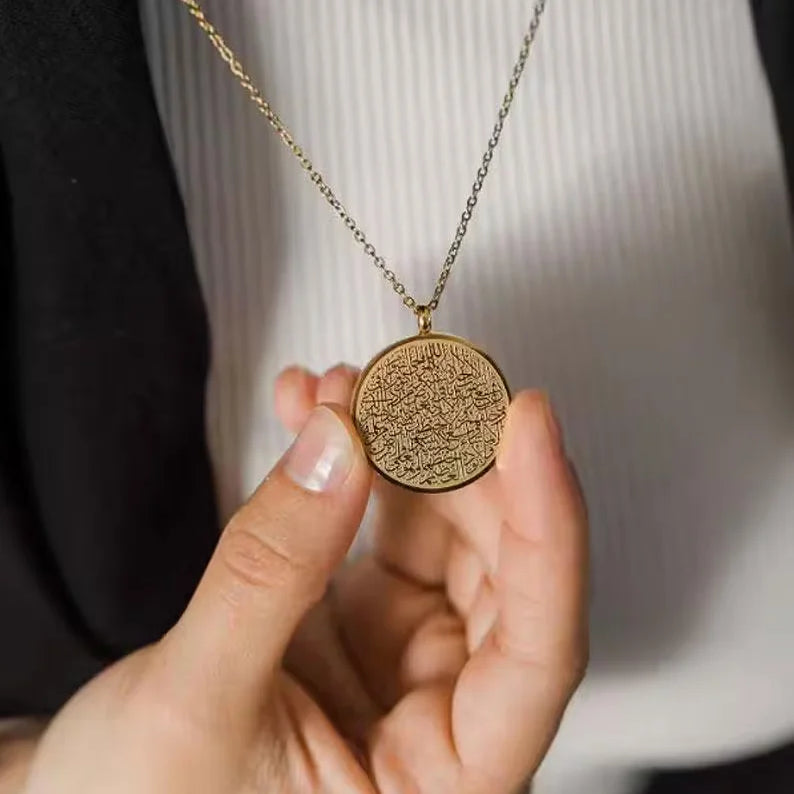 A close-up of a person holding a round gold pendant necklace with intricate Arabic calligraphy, wearing a white ribbed shirt and a black blazer.