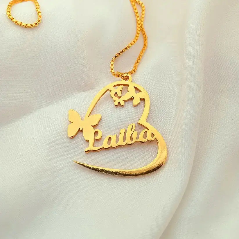 A gold nameplate necklace featuring a heart-shaped outline with the name 'Laiba' and butterfly motifs, displayed on a soft white fabric background.