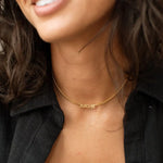 A close-up lifestyle shot of a woman wearing a delicate gold chain necklace with the name 'ARCHIE' engraved in small capital letters on the front.
