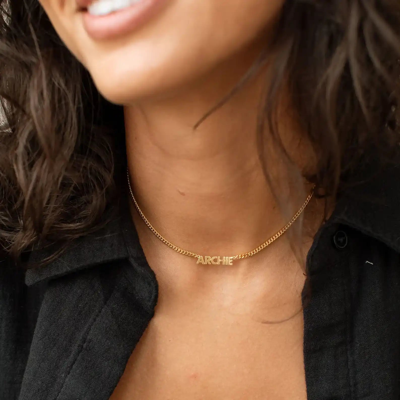 A close-up lifestyle shot of a woman wearing a delicate gold chain necklace with the name 'ARCHIE' engraved in small capital letters on the front.