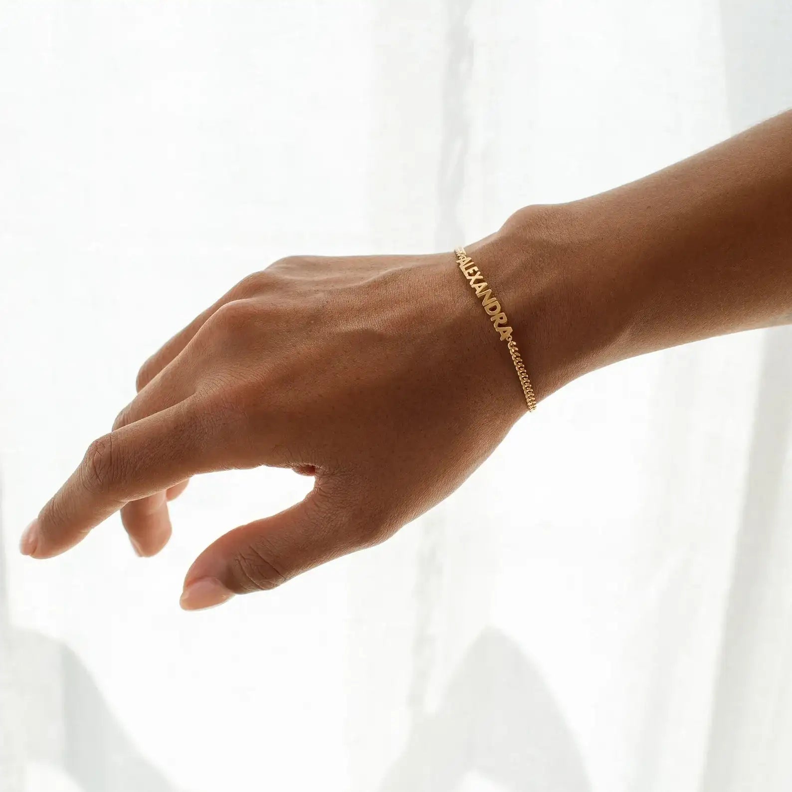 A gold chain bracelet with repeating names spelling 'ALEXANDRA' worn on a person's wrist, shown against a bright white curtain background.