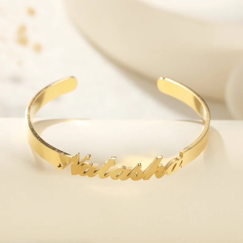 Custom name bangle bracelet with flowing script lettering across the front of a sleek open cuff design, shown in a golden finish.