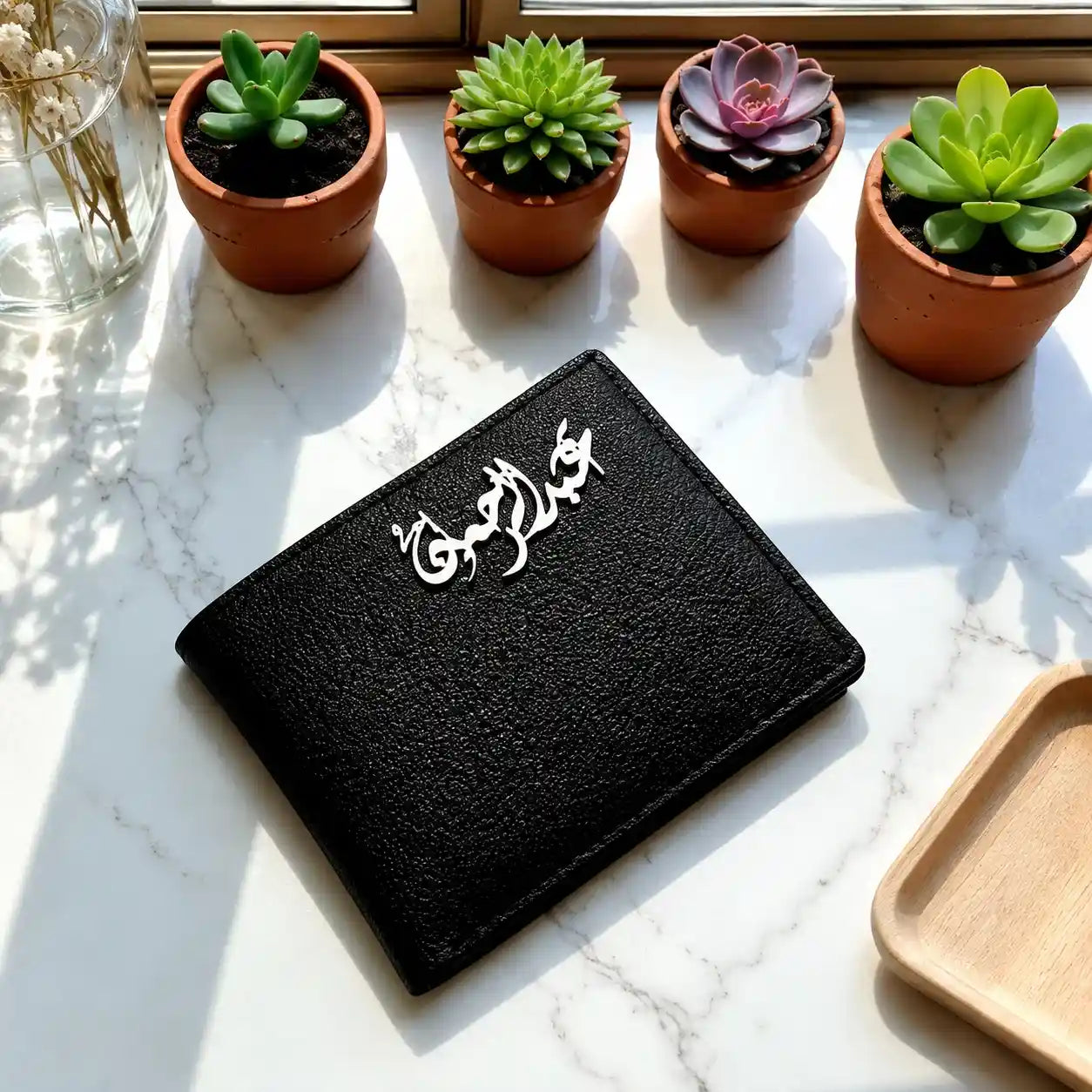 Personalized black leather wallet with custom silver Arabic calligraphy emblem displayed on a white marble countertop next to potted succulents.