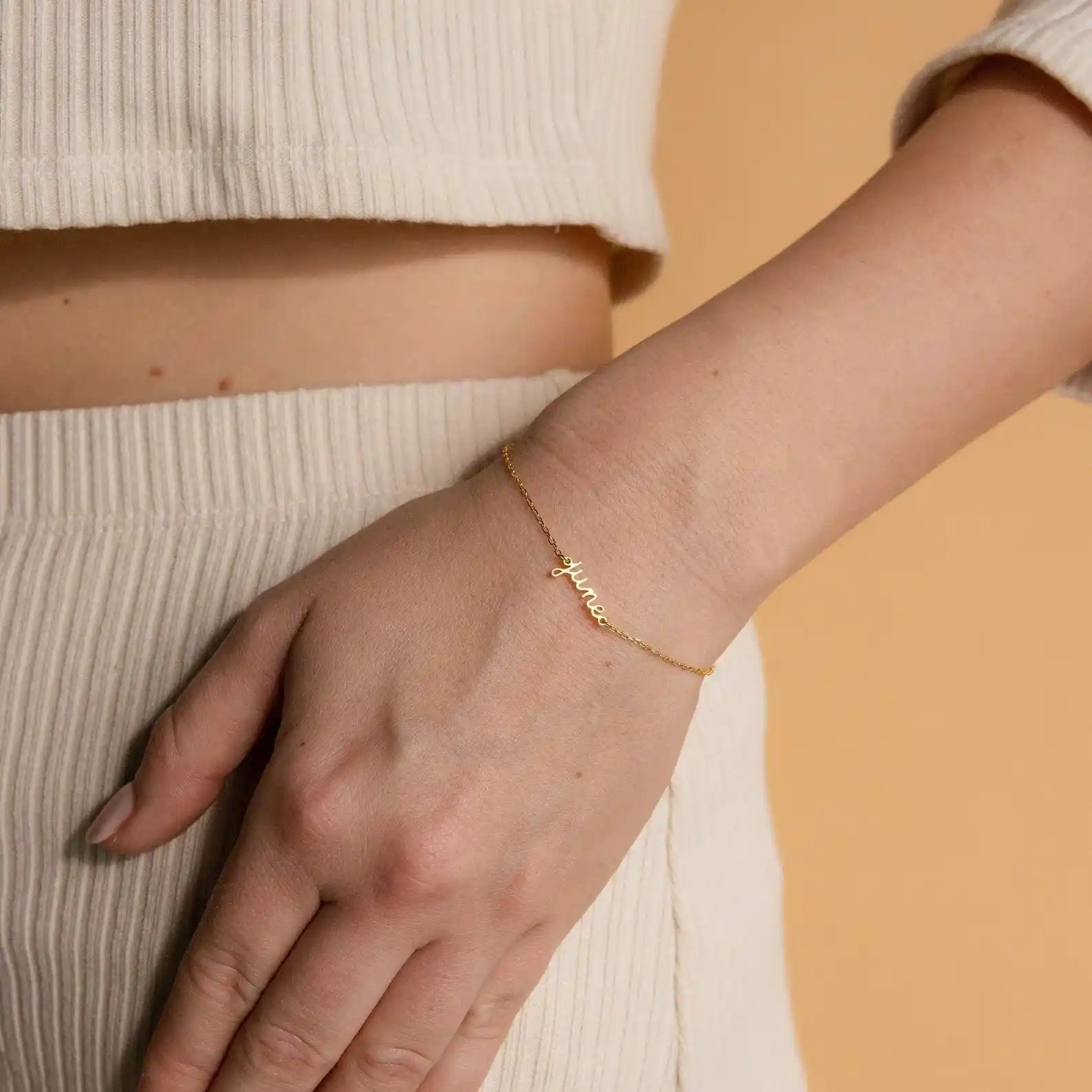 Woman wearing fine gold chain bracelet with june cursive script nameplate on wrist paired with cream ribbed outfit