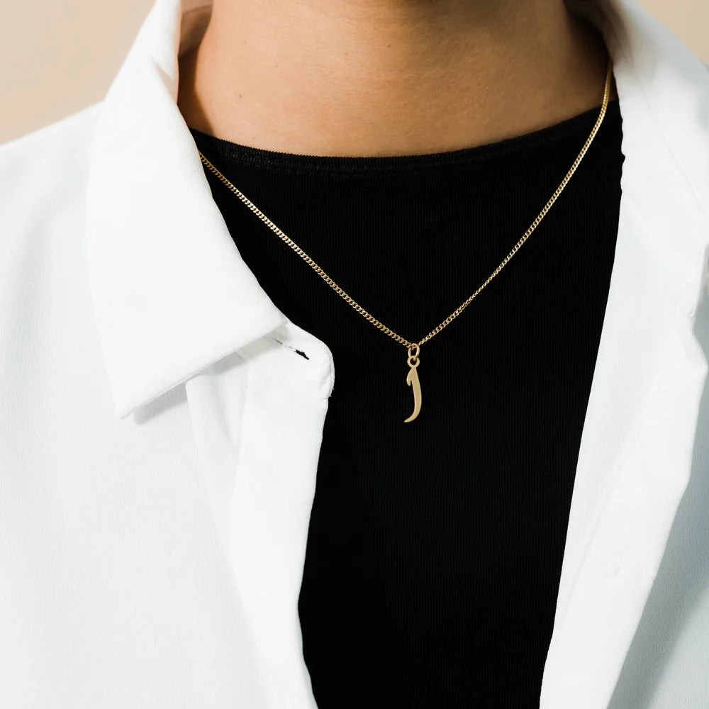 Close-up of a gold Arabic initial letter pendant on a fine link chain worn over a black top and white open shirt.