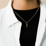 Close-up of a gold Arabic initial letter pendant on a fine link chain worn over a black top and white open shirt.