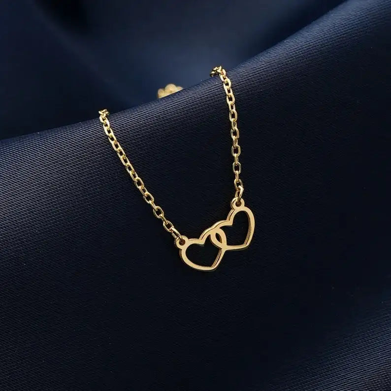 A delicate gold chain necklace featuring two interlocking open heart pendants, displayed on a dark navy blue textured fabric background.