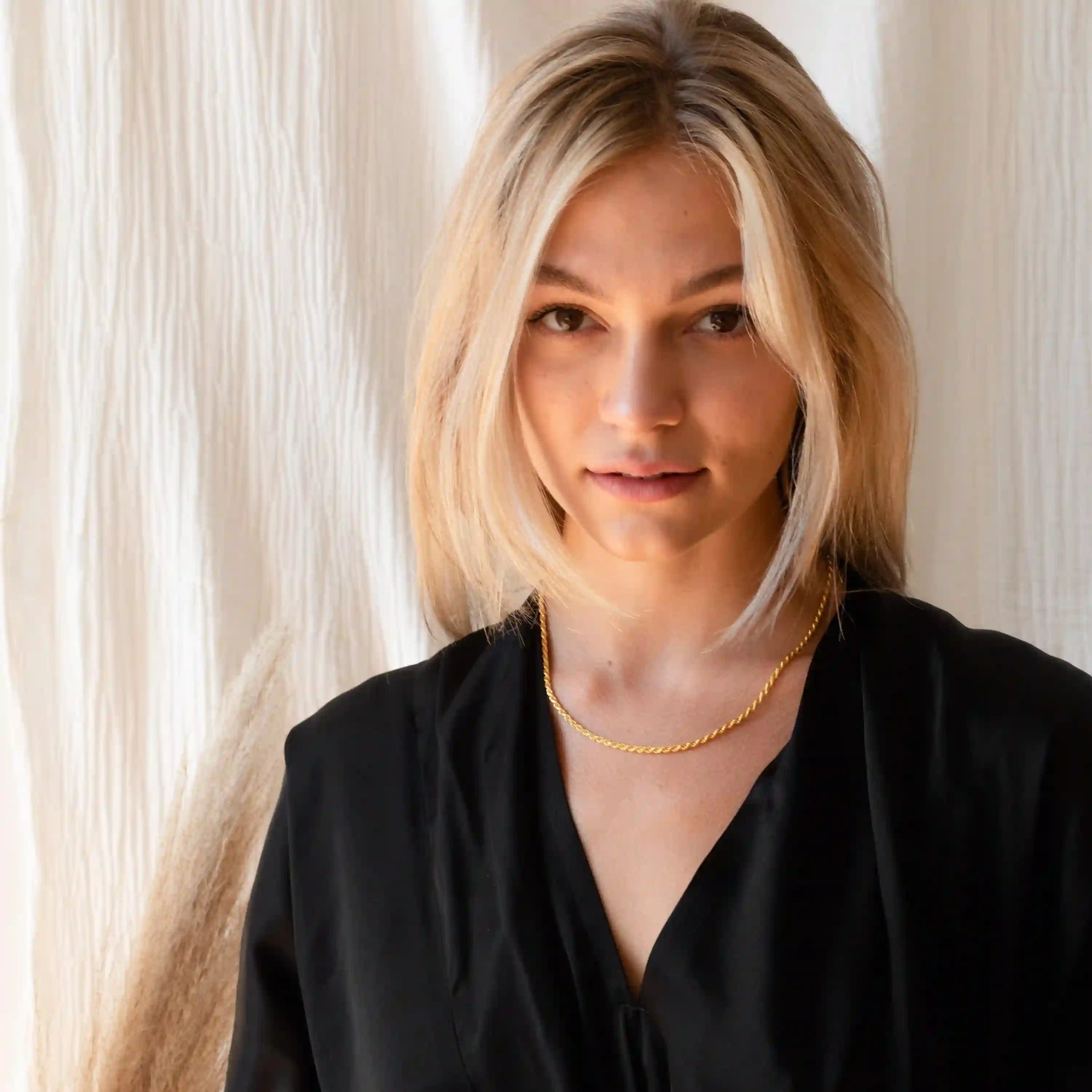 Blonde woman wearing thin gold twisted rope chain necklace against neutral fabric backdrop