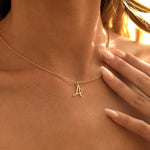 A close-up of a model's neck and chest wearing a delicate gold chain necklace with a small cursive letter 'A' pendant.