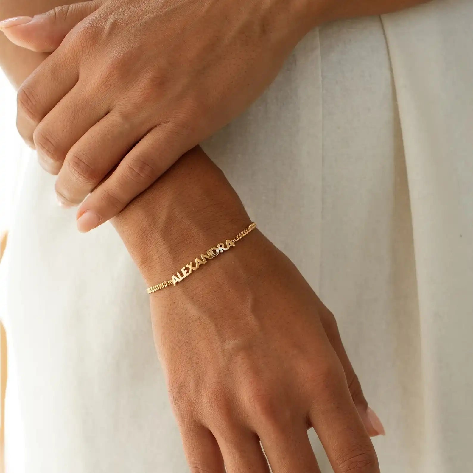 Gold bracelet with 'ALEXANDER' on a wrist against a neutral background
