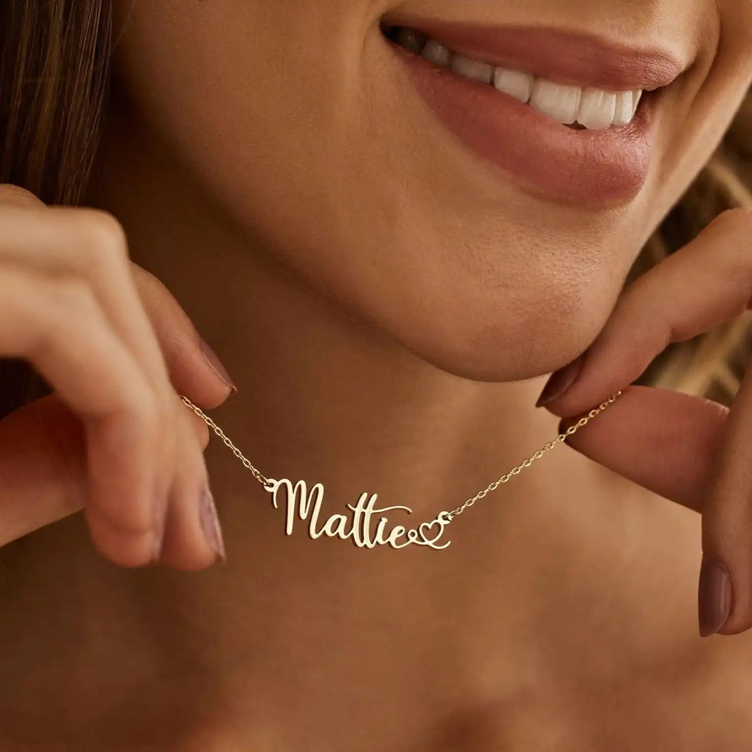 A close-up lifestyle shot of a woman wearing a delicate gold script necklace that reads 'Mattie' with a small heart at the end, held gently by her hands.