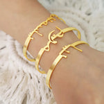 Three gold-tone Arabic calligraphy cuff bracelets worn on a person's wrist, shown against a soft white textured background.