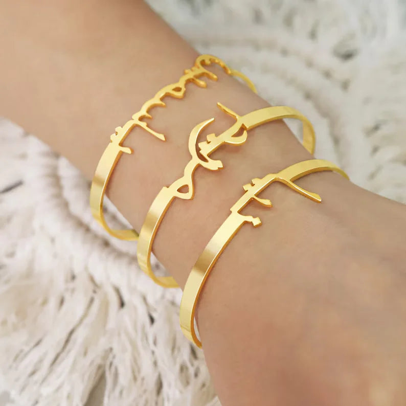 Three gold-tone Arabic calligraphy cuff bracelets worn on a person's wrist, shown against a soft white textured background.