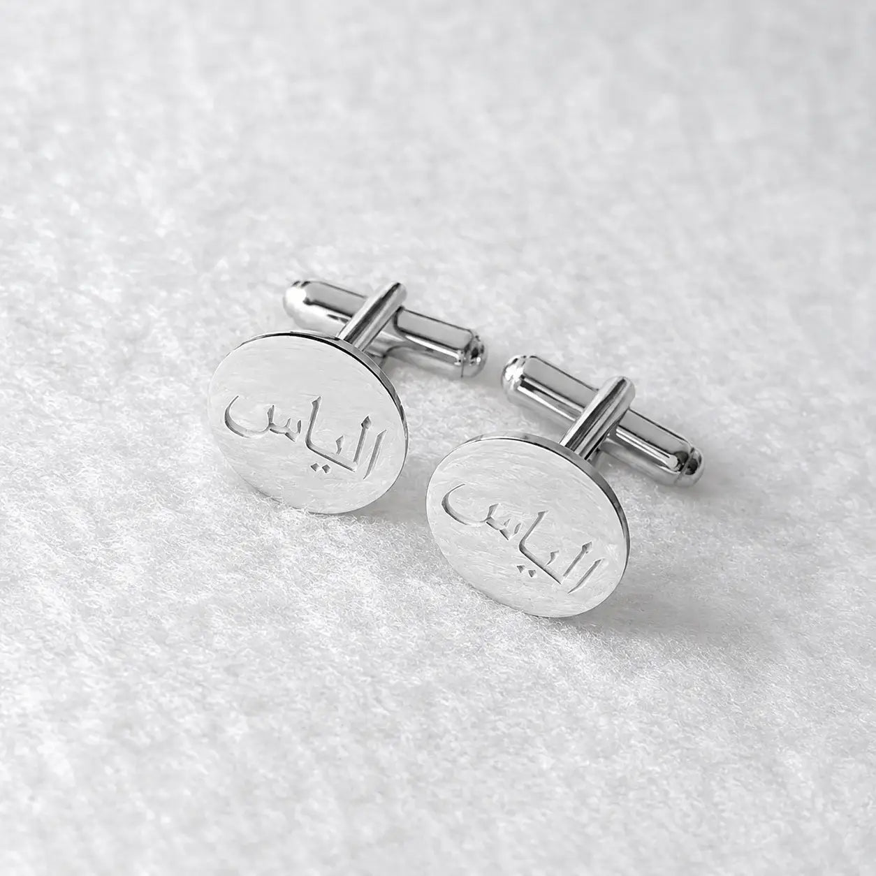 A pair of silver oval cufflinks engraved with Arabic calligraphy, displayed on a textured white fabric background.