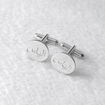 A pair of silver oval cufflinks engraved with Arabic calligraphy, displayed on a textured white fabric background.