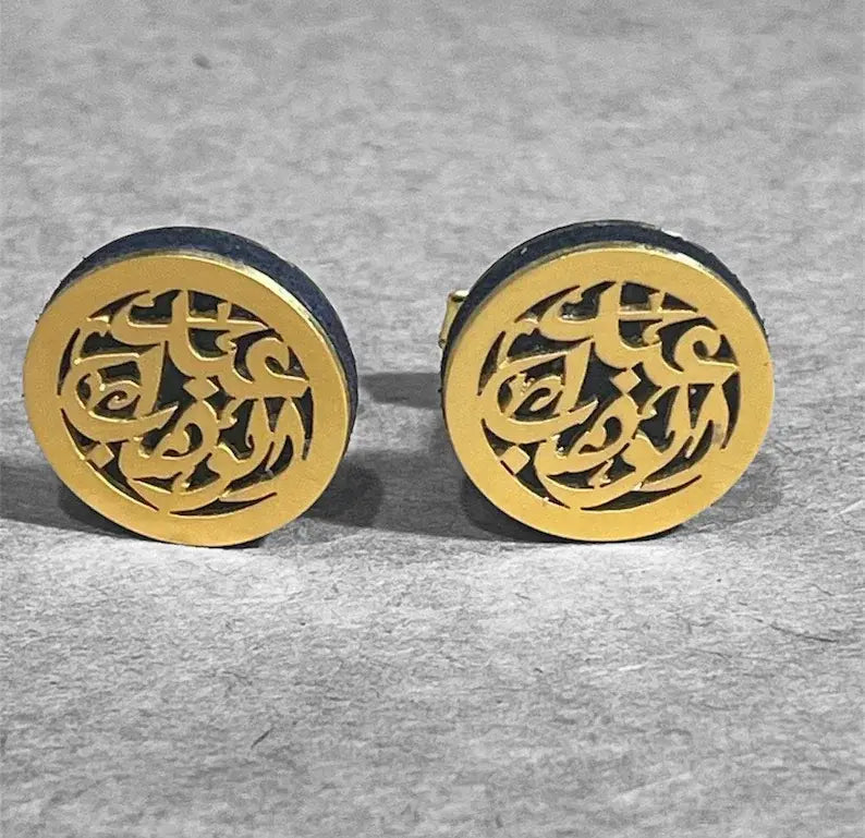 A pair of round gold-tone cufflinks with intricate cutout Arabic calligraphy patterns, set against a textured gray background.