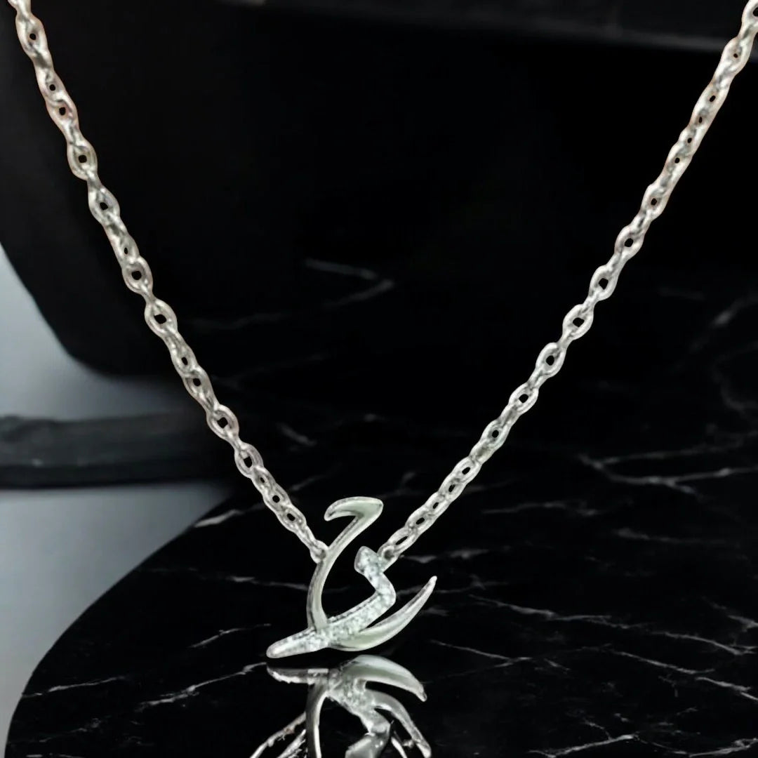 Silver Arabic calligraphy initial pendant encrusted with pave crystals, displayed on a fine cable chain over a reflective black marble surface.