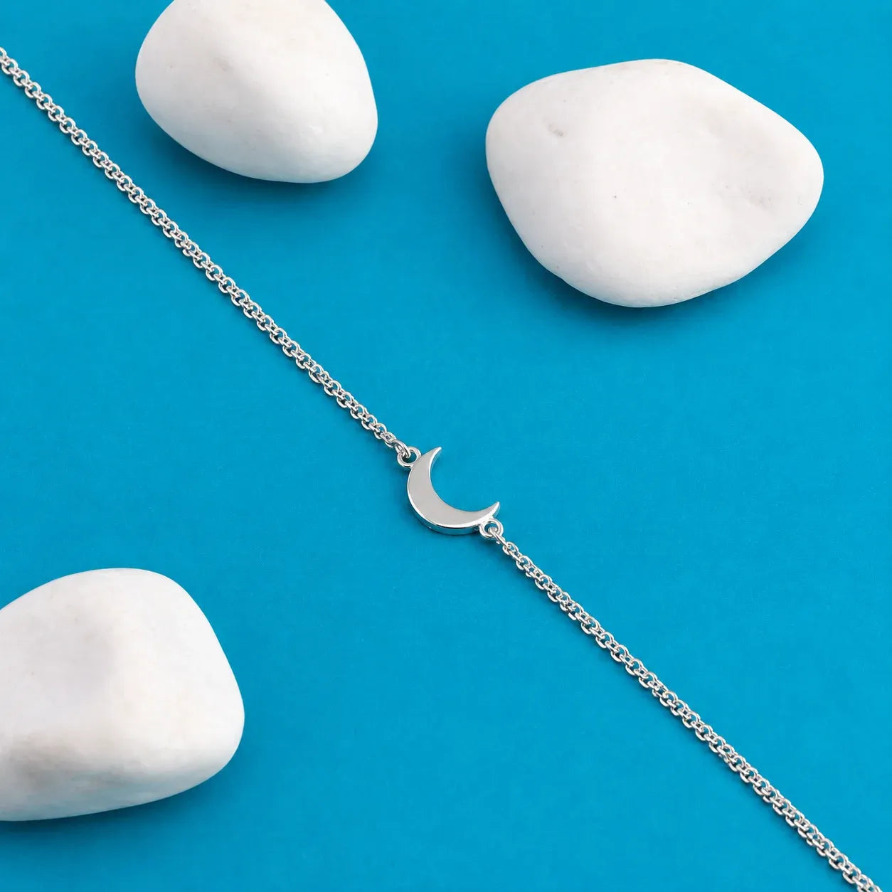 A delicate silver chain bracelet with a small crescent moon charm, laid diagonally on a bright blue background with three smooth white stones.