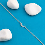 A delicate silver chain bracelet with a small crescent moon charm, laid diagonally on a bright blue background with three smooth white stones.