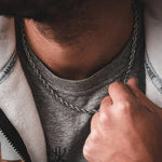 A close-up of a man wearing a silver twisted rope chain necklace over a gray t-shirt and white hoodie.
