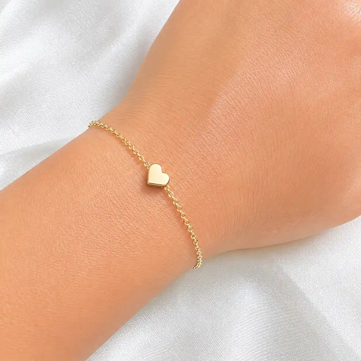 A delicate gold chain bracelet with a small flat heart charm, shown worn on a person's wrist against a soft white fabric background.