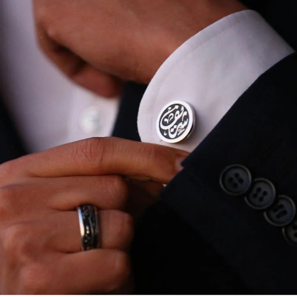 Man wearing white dress shirt and black suit jacket displaying round silver cufflink with custom black Arabic calligraphy insert