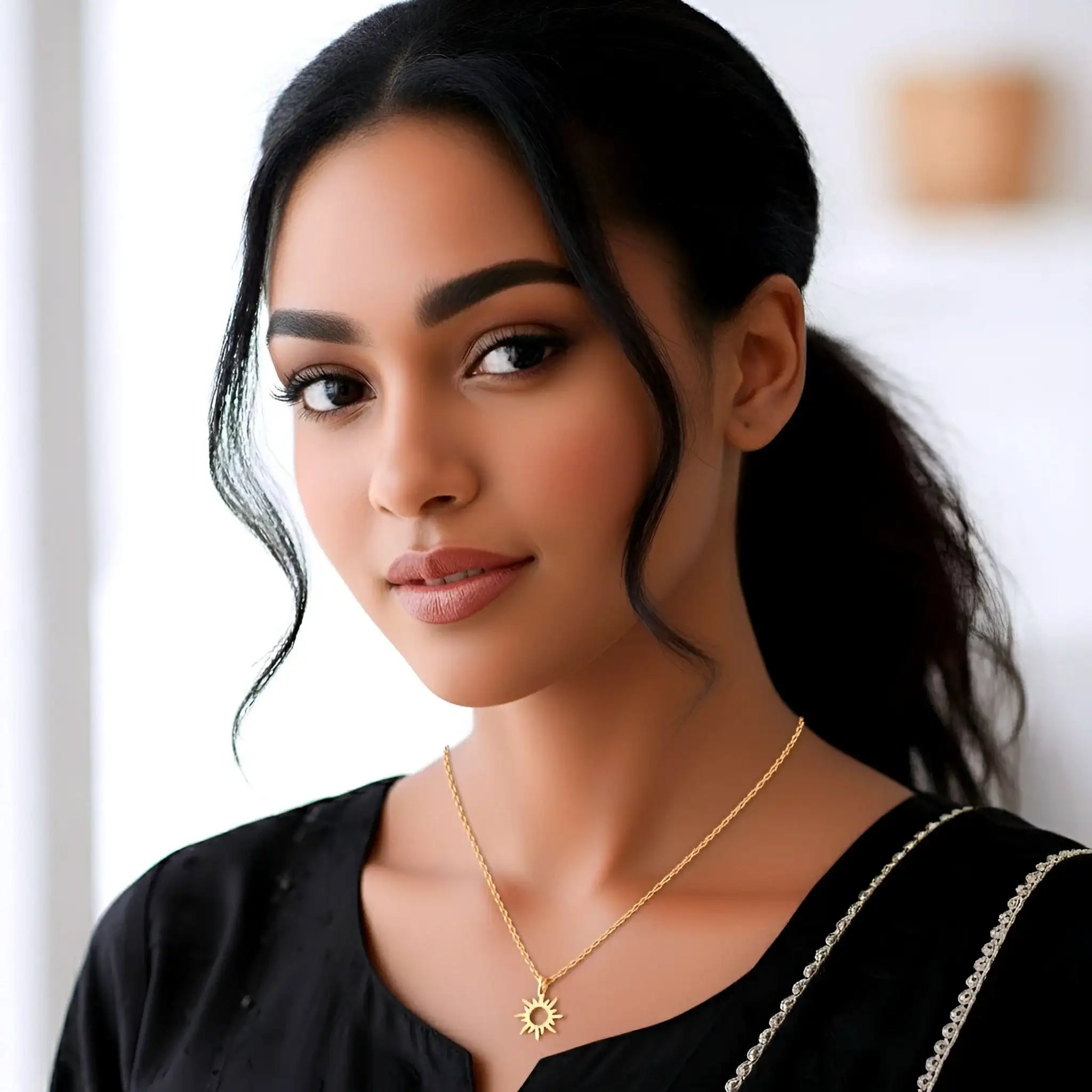 A woman with dark hair tied back wears a delicate gold chain necklace with a small sunburst pendant, paired with a black top. The background is a softly blurred light interior.