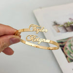 A hand holds a gold-plated bangle bracelet with the names 'Taylor' and 'Reign' engraved in cursive script, set against a softly blurred background.