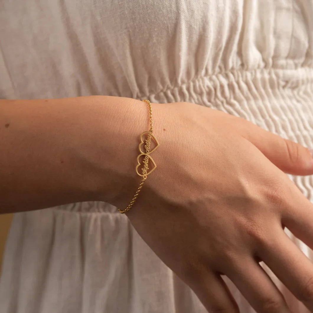 A delicate gold chain bracelet with three interlocking open heart charms, shown worn on a person's wrist against a soft beige dress.