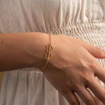 A delicate gold chain bracelet with three interlocking open heart charms, shown worn on a person's wrist against a soft beige dress.