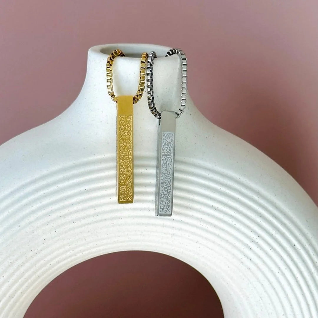 Two vertical, engraved bar pendants on matching gold and silver chains draped over a white ribbed ceramic vase against a pink background.