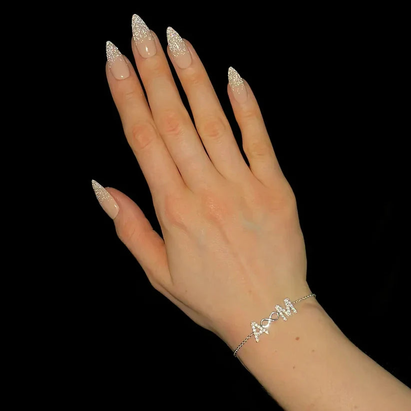 A hand with long, glittery nude-and-white manicured nails wearing a delicate silver chain bracelet with a bow and arrow charm spelling 'A&M', set against a solid black background.